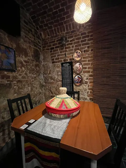 A table with a closed mesob, beside a chart of the Ethiopian alphabet on the brick wall.